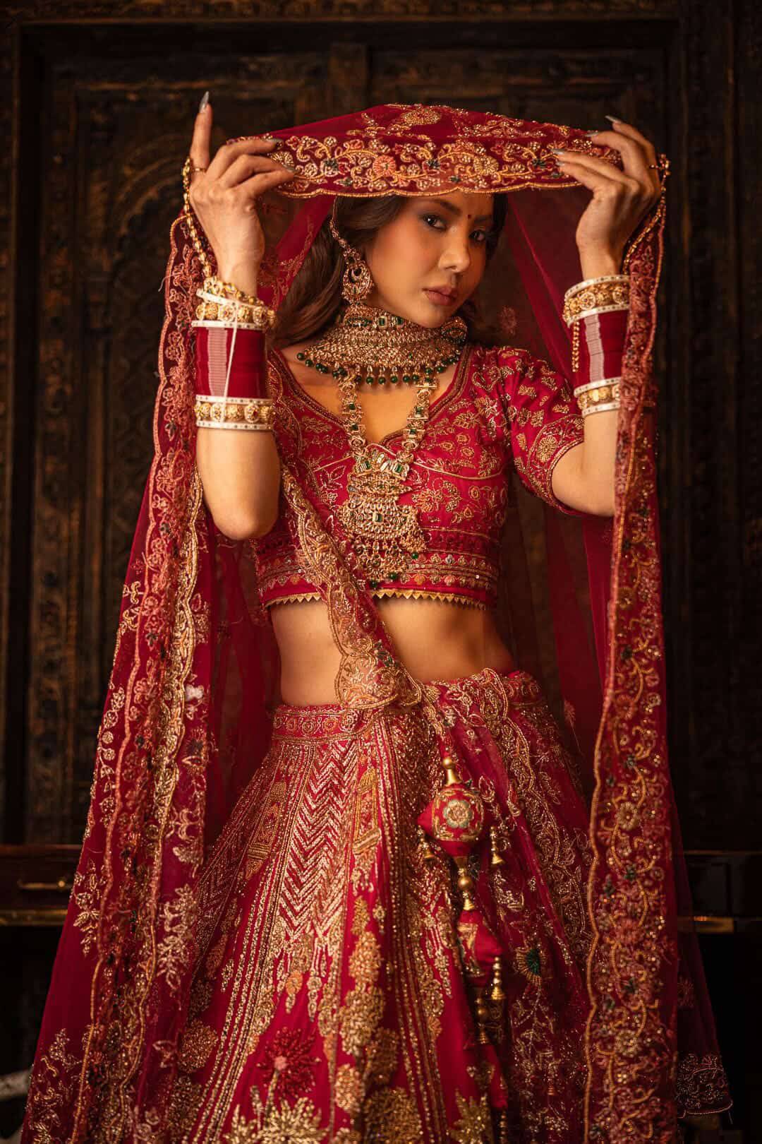 Bride in a red and gold lehenga framing her face with her embroidered dupatta, hair and makeup by Eleglam